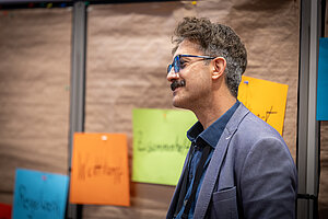 Ein Mann mit Brille und Schnurrbart steht vor einer Wand voller Haftnotizen.